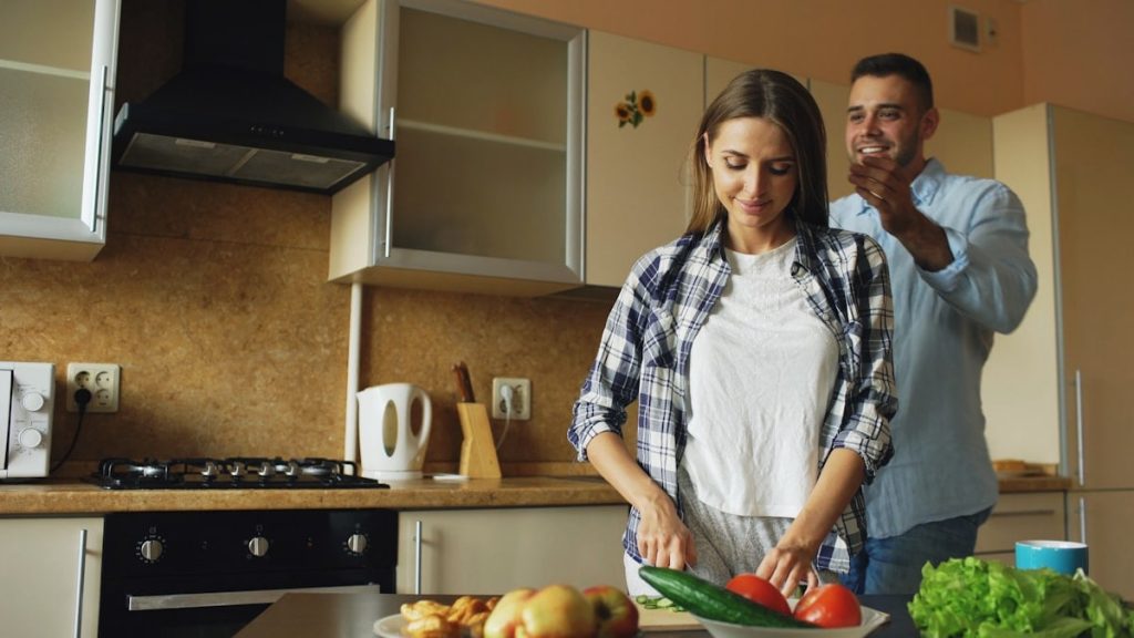 Young man covering girlfriends eyes with hands and surprising her in the kitchen at home. Loving couple cooking breakfast in the morning