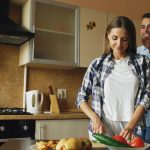 Young man covering girlfriends eyes with hands and surprising her in the kitchen at home. Loving couple cooking breakfast in the morning