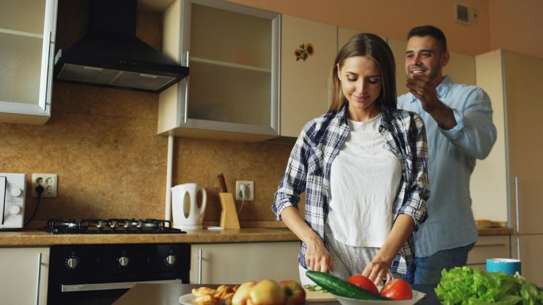 Young man covering girlfriends eyes with hands and surprising her in the kitchen at home. Loving couple cooking breakfast in the morning